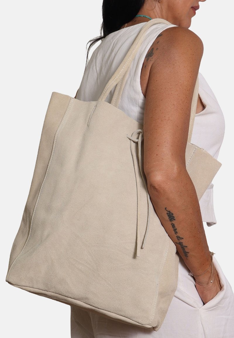 Borsa tote in suede beige con due manici lunghi, forma geometrica e dettaglio con chiusura a laccetto. Texture liscia, design minimalista.