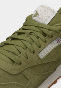 Nærbillede af en olivengrøn Reebok-sneakers med strukturerede snørebånd, ruskindss detaljer og logo med et Union Jack-flag på sidepanelet.