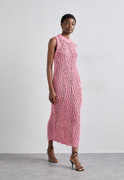 Rochie maxi tricotată roz cu modele unduitoare, fără mâneci și cu guler înalt, completată de sandale negre cu toc înalt.