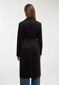 Cappotto nero in lana con vita cinturata, maniche lunghe e colletto a revers, caratterizzato da una texture liscia e una silhouette di lunghezza media.