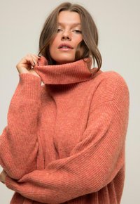 Maglione rosa a collo alto con texture a coste, vestibilità oversize e spalle scese. Il materiale appare morbido e caldo, con un motivo sottile.