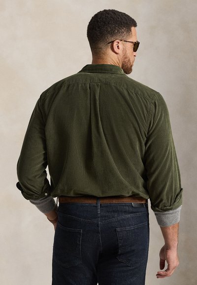 Homme aux cheveux courts portant des lunettes de soleil, une chemise en velours côtelé verte avec manches retroussées, des poignets gris et un jean bleu foncé vu de dos.