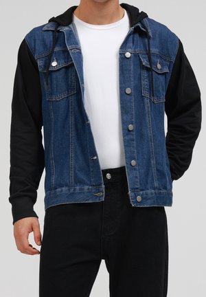 Mand iført blå denimjakke med sorte hættetrøjeærmer og en hvid t-shirt, kombineret med sorte bukser, stående med hænder nær lommerne.