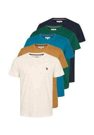 Mężczyzna uśmiechający się z rękami w kieszeniach, ubrany w kremowy t-shirt, brązowe spodnie sztruksowe i brązowe skórzane buty na jednolitym tle.