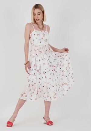 Witte maxi-jurk met bloemenpatroon, met een gelaagd ontwerp, dunne bandjes en een vloeiende silhouet. Gecombineerd met rode sandalen met doorzichtige hakken.