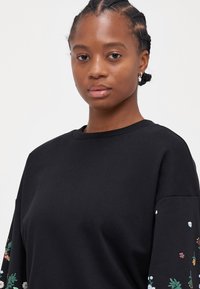 Melna džemperis ar apaļu kaklu un pārāk lielu piegriezumu, kurā uz piedurknēm ir ziedu izšuvumi dažādās krāsās. Mīksta auduma tekstūra.