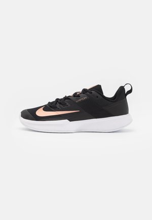 Zapatilla deportiva negra con suela blanca, logo Nike en oro rosa y marca "Vapor" en el lateral, cordones planos y tira de agarre en el talón.