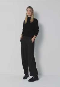 Zwarte trui met hoge hals, gecombineerd met grijze wijde pantalon met knoopaccenten. Model staat met de handen in de zakken. Zwarte enkellaarsjes.