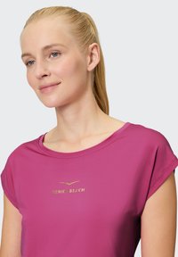 Fuchsienfarbige Kurzarmbluse mit lockerem Schnitt, vorne mit einem goldfarbenen "Venice Beach"-Logo; glatte Textur und schlichtes Design.