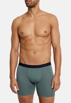 Muskulöser Mann in grünen Boxershorts mit schwarzem Bund, stehend vor einfachem weißem Hintergrund, Torso sichtbar.