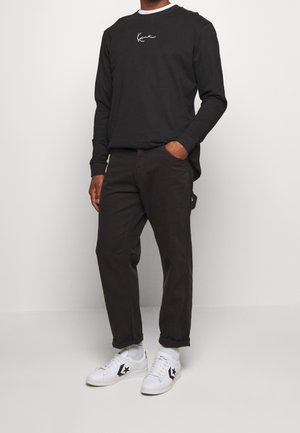 Black long-sleeve shirt with white logo, paired with dark brown pants and white sneakers featuring black star accents. Smooth fabric texture.