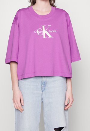 Osoba ubrana w luźną, fioletową koszulkę Calvin Klein Jeans zestawioną z jasnoniebieskimi, podartymi dżinsami na jednolitym tle.