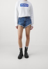 Maglione bianco lavorato a maglia con logo blu "HUGO", pantaloni corti in denim con orli sfilacciati e stivali neri alla caviglia con chiusura lampo e suola spessa.