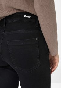 Zwarte spijkerbroek van denim met een gladde textuur, achterzakken met een subtiele stikselafwerking en een wit "Brax" logo op de tailleband.