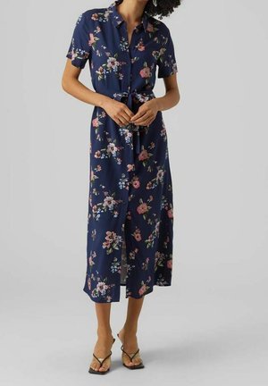 Kobieta ubrana w granatową kwiecistą sukienkę midi z krótkimi rękawami, kołnierzykiem, zapinaną na guziki z przodu i zawiązaną w talii, w parze z czarnymi sandałami na paski.