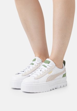 White sneakers with a textured platform sole, featuring beige suede and green accents. Laces are white; branding visible in gold.
