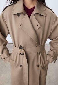 Impermeabile beige realizzato in tessuto liscio, con una chiusura doppiopetto, ampi revers e vita con cintura e fibbia rettangolare.
