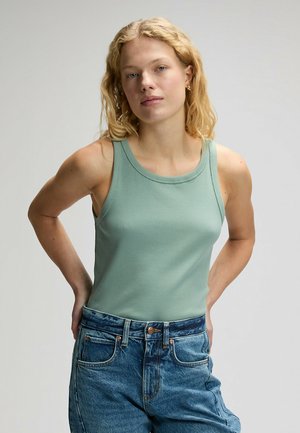 Junge Frau mit blondem Haar, die ein grünes geripptes Tanktop und blaue hoch taillierte Jeans trägt, steht mit den Händen hinter dem Rücken.