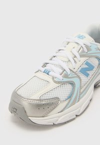 Weißer Sportschuh aus Mesh-Material, mit silbernen Akzenten und hellblauen Details. Verfügt über eine runde Zehenpartie, eine strukturierte Oberseite und ein Schnürdesign.