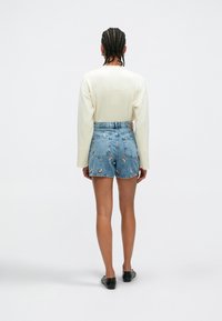 Pantalones cortos de mezclilla con bordado floral multicolor; diseño de cintura alta, dobladillo deshilachado, combinados con una blusa de manga larga color crema y zapatos planos negros.