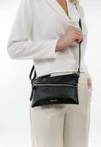 Zwarte leren schoudertas met een gestructureerde afwerking, voorzien van een ritssluiting en een verstelbare schouderband, gedragen over een wit shirt en beige broek.