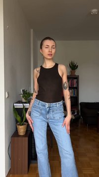 Νεαρή γυναίκα με τατουάζ, φοράει μαύρο αμάνικο τοπ και ανοιχτόχρωμο τζιν, στέκεται σε ένα σαλόνι με φυτά και ράφια βιβλίων.