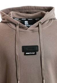 Sudadera marrón con un bolsillo delantero, cordones y dos etiquetas decorativas: una negra y en relieve, la otra con "MWM Design."