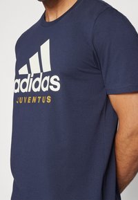 Mężczyzna w granatowej koszulce Adidas z białym logo i złotym napisem "Juventus", widoczny od ramion do talii.