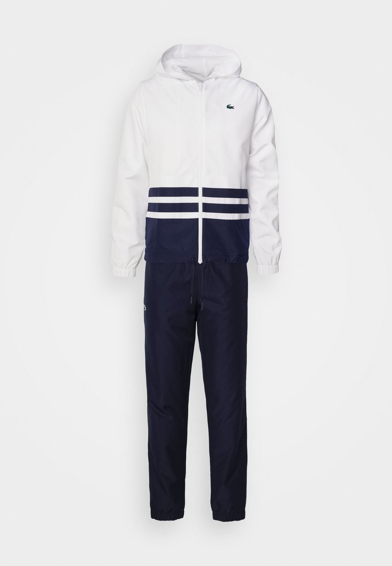 Chándal que incluye una chaqueta blanca con capucha y detalles en azul marino, adornada con tres franjas blancas; combinada con pantalones azul marino que tienen un dobladillo elástico.