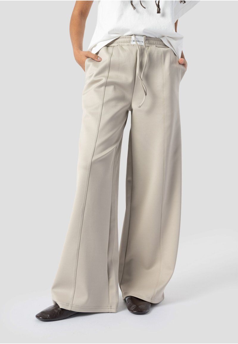 Pantalon beige à jambes larges avec coutures frontales et taille élastique, porté avec un haut blanc et des chaussures foncées à bout pointu, mains dans les poches.