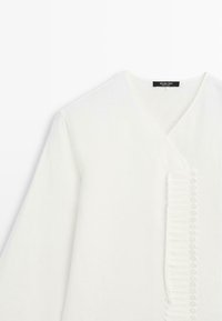 Blouse blanche à manches longues avec col en V, rangée verticale de petits boutons blancs et détails plissés le long du centre avant.
