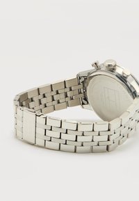 Ασημένιο μεταλλικό βραχιόλι ρολογιού με κούμπωμα, μερικώς ανοιχτό, που δείχνει την πίσω πλευρά της στρογγυλής κάσας ρολογιού με κουμπιά ρύθμισης.