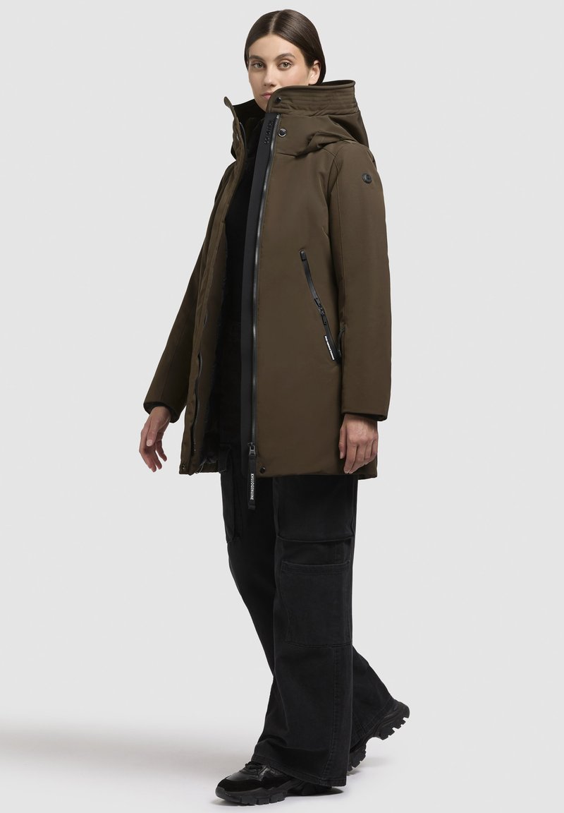 Mujer vestida con un abrigo acolchado de oliva oscuro con capucha, pantalones cargo negros y zapatillas negras, de pie en una pose neutra sobre un fondo claro.