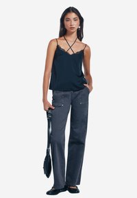 Canotta della Marina con pizzo, abbinata a pantaloni larghi grigi con tasche borchiate. Modella che tiene una borsa nera, indossando scarpe basse.