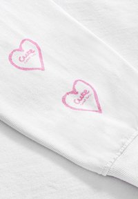 Biała bawełniana tkanina z dwoma różowymi wzorami serc z napisem "Cute" w środku, umiejscowionymi na końcu rękawa. Widoczne detale faktury.