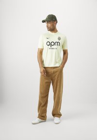 Svetložltý športový dres s čiernym logom, skombinovaný s hnedými kordovým nohavicami a bielymi teniskami. Model má na sebe tmavozelenú čiapku.