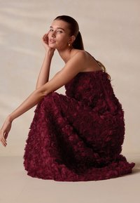 Bordeauxrode bloemenjurk gemaakt van gelaagde stoffen rozen, getailleerd lijfje met dunne bandjes, vloeiende rok met gestructureerde gerafelde details.