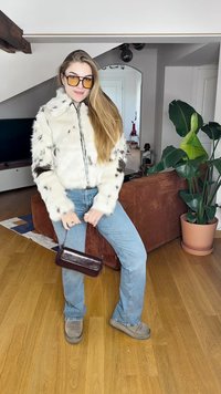 Faux fur jacket in off-white with brown spots, front zipper, worn with light blue jeans and brown boots, holding a small brown handbag.