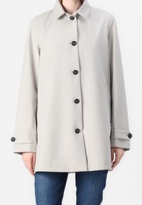 Lichtbeige lange mouw knoopjas met zwarte knopen, gedragen over een blauwe spijkerbroek, getoond van de nek tot halverwege de dij.