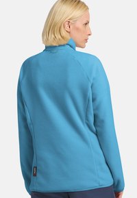 Giacca in pile azzurro chiaro con colletto alto, maniche raglan e una sottile texture. Presenta un piccolo etichetta nera del marchio sul lato.