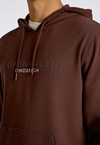 Brun hoodie i mjukt tyg, med en känguruficka och en iögonfallande tryck "COPENHAGEN LINDENBERGH" över bröstet.