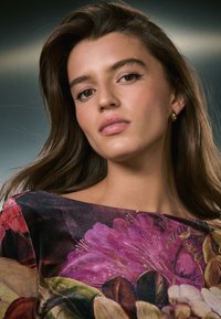 Model mit langem, dunklem Haar, die ein Oberteil mit floralem Muster in lebendigen Pink- und Lilatönen trägt, kombiniert mit goldenen Creolen.