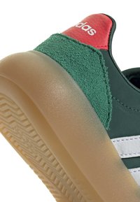 Close-up of Adidas sneaker heel with green suede, red leather detail, white stripes, and translucent rubber sole.