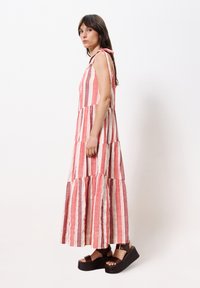 Langes, gestuftes Kleid mit roten und cremefarbenen vertikalen Streifen, aus leichtem Stoff gefertigt. Verfügt über Schulterbänder und eine fließende Silhouette, kombiniert mit Plateausandalen.