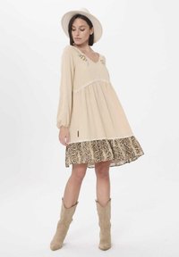 Beige jurk met lange mouwen, v-hals, bloemenborduursel, kanten afwerking en ruffle zoom in slangenprint. Gecombineerd met beige enkelboots en een brede hoed.