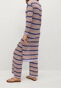 Ensemble en tricot à motifs zigzag violet et beige, avec des manches longues et un pantalon à jambes larges. Porté avec des sandales brunes simples.