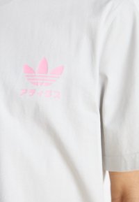 Vit bomullströja med en rosa stiliserad logotyp med tre blad och ränder, som är placerad på vänster bröstområde.