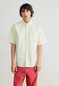 Kortermet, lys gul skjorte med knapper og brystlomme, laget av glatt stoff, sammen med røde bukser. Enkel design, uten mønstre.