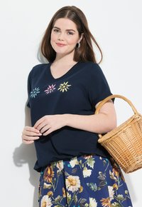 Camiseta de cuello en V de color marino con tres bordados florales coloridos en rosa, azul y amarillo. Llevada con una falda floral y sosteniendo una cesta de mimbre.