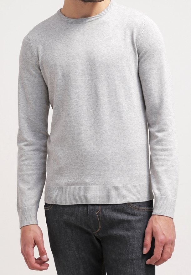 Homme portant un pull gris clair à manches longues col rond et un jean foncé, debout devant un fond blanc uni.